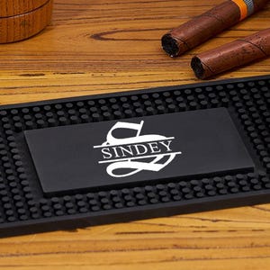 May include: A rectangular black rubber bar mat with a textured nub surface and a central smooth panel featuring the white printed text "SINDEY" beneath a stylized letter "S". The mat is positioned horizontally on a wooden surface.