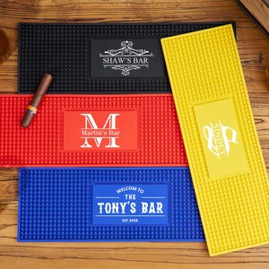 May include: A collection of four rectangular rubber bar mats in black, red, blue, and yellow. The black mat features the text 'SHAW'S BAR', the red mat displays 'Martin's Bar' with a large 'M', the blue mat says 'WELCOM TO THE TONY'S BAR EST.2028', and the yellow mat shows 'SINDEY'.