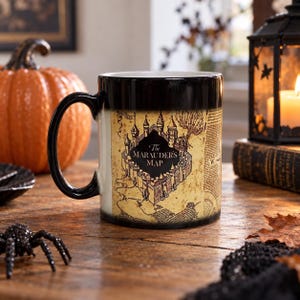 May include: A black ceramic mug featuring a tan, parchment-style graphic of The Marauder's Map with a central illustration of a castle. The design includes intricate line work, text, and compass markings printed across the exterior surface.