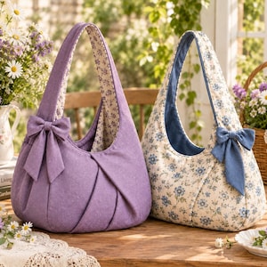 Peut inclure: Deux sacs à main de style hobo avec une bandoulière unique et un nœud décoratif sur le côté. Un sac est lavande uni avec une doublure intérieure florale, tandis que l'autre présente un imprimé floral bleu sur fond crème avec un nœud bleu uni.