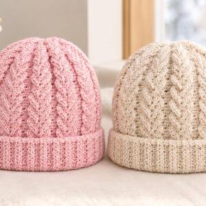 May include: Two knit beanies, one in light pink and one in cream, featuring a vertical cable knit pattern on the crown and a ribbed cuff at the base. The hats are constructed with a textured yarn, showcasing a consistent stitch design throughout.