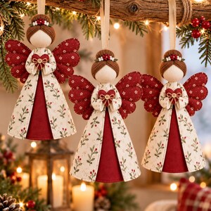 May include: Three hanging angel ornaments featuring cream-colored fabric dresses with a red berry and green leaf pattern, deep red fabric wings, and brown yarn hair. Each angel holds a small red fabric heart with a central button detail.