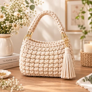 Puede incluir: Un bolso de ganchillo color crema con un patrón de tejido grueso y texturizado, un asa redondeada y herrajes de metal dorado. Una borla larga de cordón a juego cuelga de un lado del bolso, sujeta por una anilla de metal dorado.