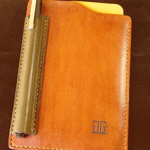 May include: A tan leather notebook cover featuring a dark olive green pen loop attached to the left side, containing a black pen with a silver clip. A yellow card is tucked into the top, and a small embossed logo is visible on the front cover.