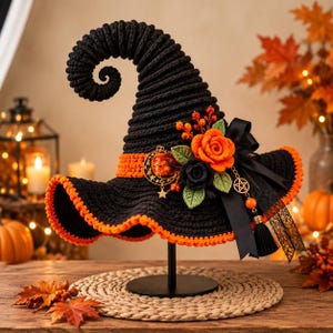 May include: A black crocheted witch hat with a curled tip and an orange brim edge. It features an orange band, a black ribbon bow, orange and black crocheted roses, green leaves, and gold-toned charms including a crescent moon and a pentagram.