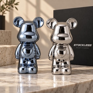 Op de afbeelding: Twee beervormige beeldjes met een glanzende, reflecterende afwerking. De ene is metallic blauw en de andere zilverkleurig. Elk figuur heeft een opvallende hartvorm op de borst en ronde oren. Een zwarte doos met de tekst STOCKLESS NEVER STANDARD is zichtbaar.