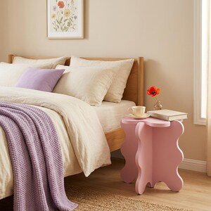 Puede incluir: Una mesa auxiliar de color rosa claro con una silueta ondulada y orgánica se encuentra junto a una cama. La mesa sostiene una taza de cerámica color crema, un pequeño florero de vidrio con una amapola roja y un libro cerrado. La cama tiene ropa beige.