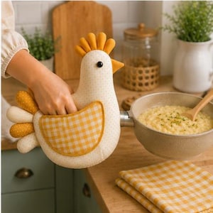 Puede incluir: Una manopla de cocina con forma de gallina hecha de tela color crema con pequeños puntos amarillos, un ala de cuadros vichy amarillos y blancos y una cresta amarilla sólida. La manopla está diseñada para cubrir el mango de una sartén.