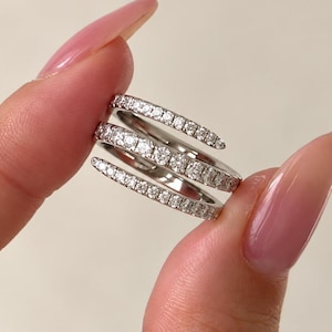 May include: A silver-toned bypass ring featuring three overlapping bands. Each band is encrusted with a row of small, clear, round-cut stones that catch the light, creating a continuous sparkling effect across the layered design of the jewelry.