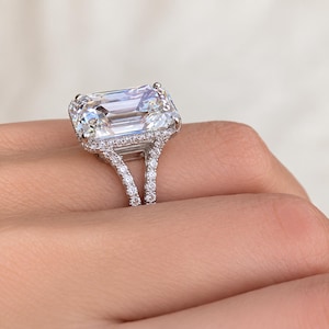 Op de afbeelding: Een zilverkleurige ring met een grote, rechthoekige, smaragdgeslepen heldere steen in een zetting met vier pootjes. De ringband is gespleten in twee delen, en zowel de halo rond de middelste steen als de gespleten band zijn ingelegd met kleine, ronde, heldere steentjes.