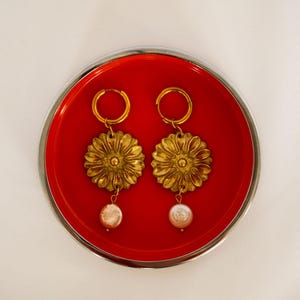 Pode incluir: Um par de brincos de argola dourados com um pendente circular em forma de flor com pétalas texturizadas e uma pequena conta redonda branca iridescente semelhante a uma pérola pendurada na parte inferior de cada berloque floral, numa bandeja vermelha.