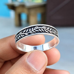 May include: A silver band ring featuring a continuous embossed leaf or vine pattern along the center. The metal has a polished finish on the edges, while the recessed areas of the botanical design are darkened to provide contrast and definition.