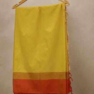 Op de afbeelding: Een felgele sari met een brede oranje rand aan de onderkant, gescheiden door een dunne goudkleurige band. De stof hangt aan een houten kledinghanger en heeft een zijkant afgewerkt met decoratieve lichtgekleurde kwastjes en rode draadaccenten.
