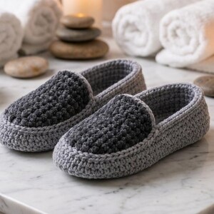 Puede incluir: Un par de pantuflas tejidas a mano con una base gris claro y un panel superior texturizado en gris carbón oscuro. Las pantuflas tienen un diseño de punta redondeada y un estilo sin cordones con una construcción gruesa y resistente.
