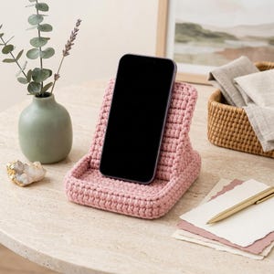 Può includere: Un supporto per telefono all'uncinetto color rosa polvere che sostiene uno smartphone nero. Il supporto presenta una struttura in filato spesso e testurizzato con uno schienale inclinato e un piccolo bordo alla base per il dispositivo.