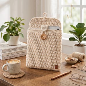 May include: A cream-colored crochet tablet sleeve featuring a textured bobble stitch pattern, a single wooden button closure, and a small rectangular brand tag at the bottom corner. The sleeve contains a silver tablet device inside.
