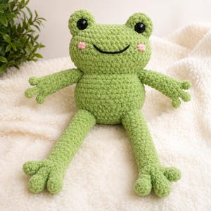 May include: A light green crochet frog plush toy with large black plastic eyes, a stitched black mouth, and small pink embroidered cheeks. The stuffed animal features long arms and legs with four distinct fingers and toes on each limb.