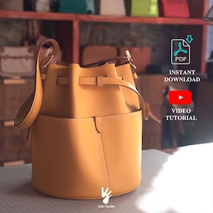 May include: A mustard yellow bucket bag with a structured cylindrical base, a top drawstring closure, and a single shoulder strap. The bag features a front pocket with a small tab closure. Text on the image includes PDF INSTANT DOWNLOAD and VIDEO TUTORIAL.