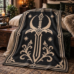 May include: A rectangular crochet blanket featuring a black background with a cream-colored central design of a sword flanked by crescent shapes and stars. The border consists of a geometric cream-colored pattern framing the entire piece.
