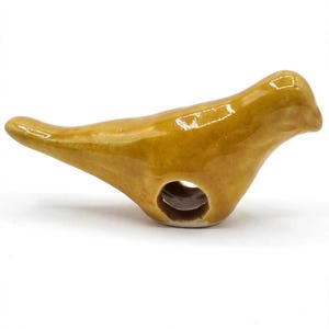 May include: A small, mustard-yellow ceramic bird whistle with a glossy, smooth finish. The bird is depicted in a horizontal, resting position with a rounded head, a pointed beak, and a tail extending backward. A circular hole is visible on the side.