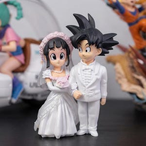 May include: A pair of anime-style figurines depicting a couple in wedding attire. The figure on the left wears a white gown with a pink floral accent and a veil. The figure on the right has black spiky hair and wears a white tuxedo with a bow tie.
