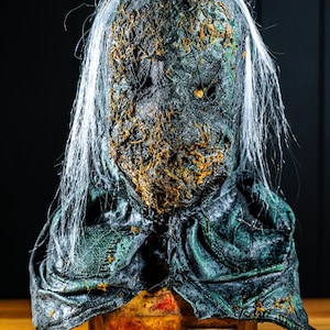 May include: A weathered, grotesque mask with a decaying appearance. The mask features a distressed, textured surface with patches of orange and green, and long white strands resembling hair. The mask rests on a rectangular base with the letters "CFX" visible.