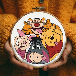 May include: A circular wooden embroidery hoop containing a cross-stitch pattern of four cartoon characters: a yellow bear in a red shirt, a pink piglet, a grey donkey, and an orange striped tiger, all stitched on a white fabric background.