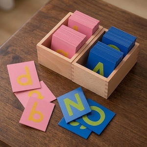 May include: Two light-colored wooden boxes containing stacks of square cards. One box holds pink cards with yellow textured letters, and the other holds blue cards with yellow textured letters. Several loose pink and blue cards are scattered in front.
