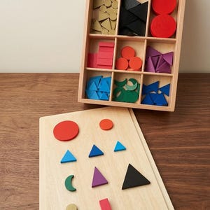 May include: A wooden sorting tray with nine compartments containing various geometric shapes in red, black, gold, pink, orange, purple, blue, and green. A matching wooden board displays a selection of these shapes arranged in a grid pattern.