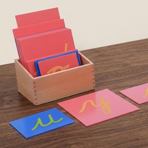 May include: A light wood box containing a stack of rectangular cards with red backgrounds and yellow cursive letters. Two additional cards are placed in front: a blue card with a green cursive letter and a red card with a yellow cursive letter.