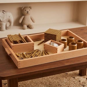 May include: A wooden tray containing a collection of maths learning materials, including a large cube of small gold beads, several wooden boxes, loose gold bead chains, and four small brown cylindrical cups arranged on a light-coloured base.