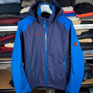 May include: A two-tone hooded jacket in dark navy and bright blue. The jacket features a full-length front zipper, two zippered side pockets, and adjustable cuffs. The Mammut brand name is printed in red on the chest, with a circular logo on the arm.