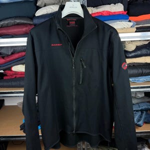 Può includere: Una giacca softshell nera con cerniera frontale integrale, colletto rialzato e una tasca verticale con cerniera sul petto. Il nome del marchio Mammut è ricamato in rosso sul petto destro, con un logo circolare rosso sulla manica sinistra.