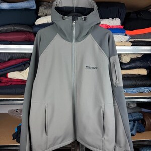 Może przedstawiać: Dwukolorowa szara kurtka typu softshell z pełnym zamkiem błyskawicznym z przodu, dołączonym kapturem z regulowanymi sznurkami oraz długimi rękawami. Na piersi znajduje się czarny napis Marmot, a na lewym rękawie kieszeń zapinana na zamek.
