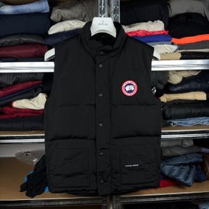 May include: A black quilted puffer vest featuring a high collar, a front snap-button closure, and two lower front pockets. A circular Canada Goose patch logo is on the left chest, and a small Canada Goose brand label is on the lower right pocket.