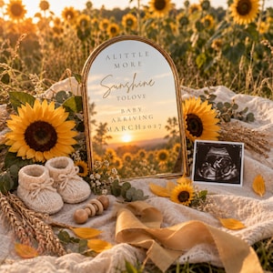 Może przedstawiać: Łukowate lustro ze złotą ramą z napisem A LITTLE MORE Sunshine TO LOVE BABY ARRIVING MARCH 2027. Lustro odbija pole słoneczników o zachodzie słońca. W zestawie znajdują się dwa beżowe dzianinowe buciki, drewniana grzechotka i zdjęcie USG.