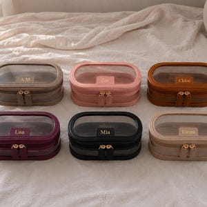 May include: Six oval-shaped jewellery cases with clear lids and gold-tone double zips. Each case features a personalised name or initials printed on the front. The cases are available in beige, pink, brown, deep purple, black, and light tan colours.