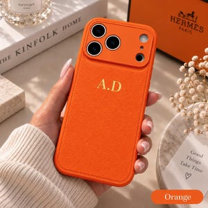 Peut inclure: Une coque de téléphone orange vif à la texture grainée, dotée d'un bloc optique surélevé avec trois découpes circulaires pour les objectifs et une ouverture pour le flash. Les initiales A.D. sont inscrites en doré au centre du dos.