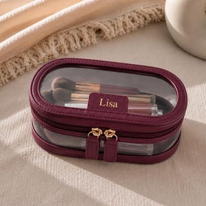 Peut inclure: Une trousse de toilette ovale bordeaux avec un panneau supérieur en plastique transparent et une fermeture éclair dorée. L'avant présente un petit écusson rectangulaire avec le nom Lisa embossé en lettres dorées. L'intérieur contient des pinceaux.