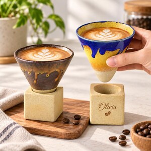 May include: Two cone-shaped ceramic coffee cups with latte art, one dark brown with a drip glaze and one yellow and blue with a drip glaze. Each cup rests on a square, light-coloured stone base. One base features the name Olivia and a coffee bean icon.
