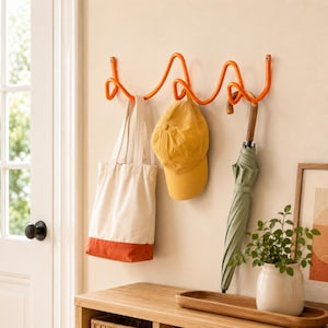 Peut inclure: Un porte-manteau mural orange vif au design métallique ondulé et continu. Suspendus aux crochets se trouvent un sac fourre-tout en toile crème avec une base orange, une casquette de baseball jaune unie et un parapluie vert sauge fermé à manche en bois.