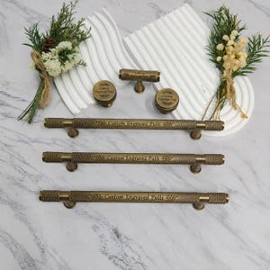 Op de afbeelding: Een collectie kastbeslag van antiek messing, bestaande uit drie lange stanggrepen, één T-greep en twee ronde knoppen. Elk stuk heeft een getextureerde gekartelde afwerking en is gegraveerd met de tekst Custom Engraved Pulls en bladaccenten.