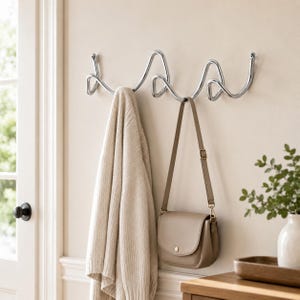Pode incluir: Um cabide de parede feito de metal cromado polido com um design ondulado contínuo. O cabide possui vários ganchos curvos para pendurar itens, com um suéter de malha bege e uma bolsa transversal de couro castanho pendurados na estrutura.
