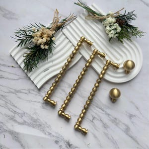 Op de afbeelding: Drie goudkleurige metalen kastgrepen met een kralentextuur en twee bijpassende ronde knoppen. Het beslag is gerangschikt naast twee witte, gewelfde, geribbelde decoratieve dienbladen, elk versierd met een klein boeket gedroogde witte bloemen en groen.