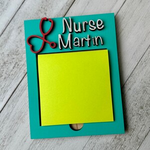 May include: A teal wooden notepad holder with a red stethoscope design and the name "Nurse Martin" in white lettering. A bright yellow sticky note pad is inside the holder.