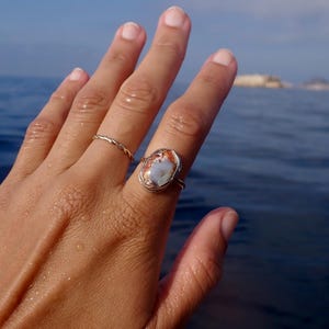 Può includere: Un anello in oro con una grande pietra ovale multicolore caratterizzata da motivi vorticosi arancioni, bianchi e marroni, montata su una semplice fascia metallica, accompagnata da un secondo anello in oro più sottile e intrecciato.