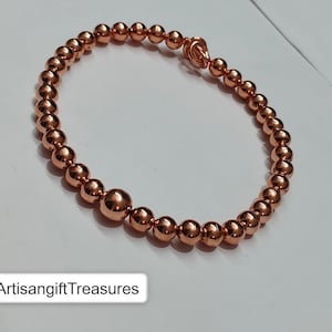 May include: A circular bracelet composed of polished copper-colored spherical beads of varying sizes, featuring a larger bead near the bottom and a small knot-like clasp mechanism at the top. The text ArtisangiftTreasures is visible.