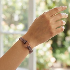 Könnte beinhalten: Ein kupferfarbenes Armband mit einem komplizierten Prägemuster auf dem Band. Das Armband wird mit einem kreisförmigen Ringverschluss und einem gedrehten Metalldetail gesichert und weist ein metallisches Finish mit dunklen Vertiefungen auf.
