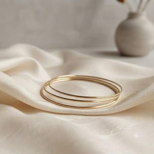 May include: A set of three thin, circular gold bangles with a smooth, polished finish. The bracelets are stacked together, showcasing a simple, continuous metal band design that maintains a consistent thickness throughout each individual piece.