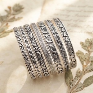 Peut inclure: Un ensemble de sept bracelets rigides en métal argenté présentant divers motifs en relief, notamment des vagues, des fleurs, des lignes texturées, des étoiles, des lunes, des tresses et de petites silhouettes de lapins empilés.